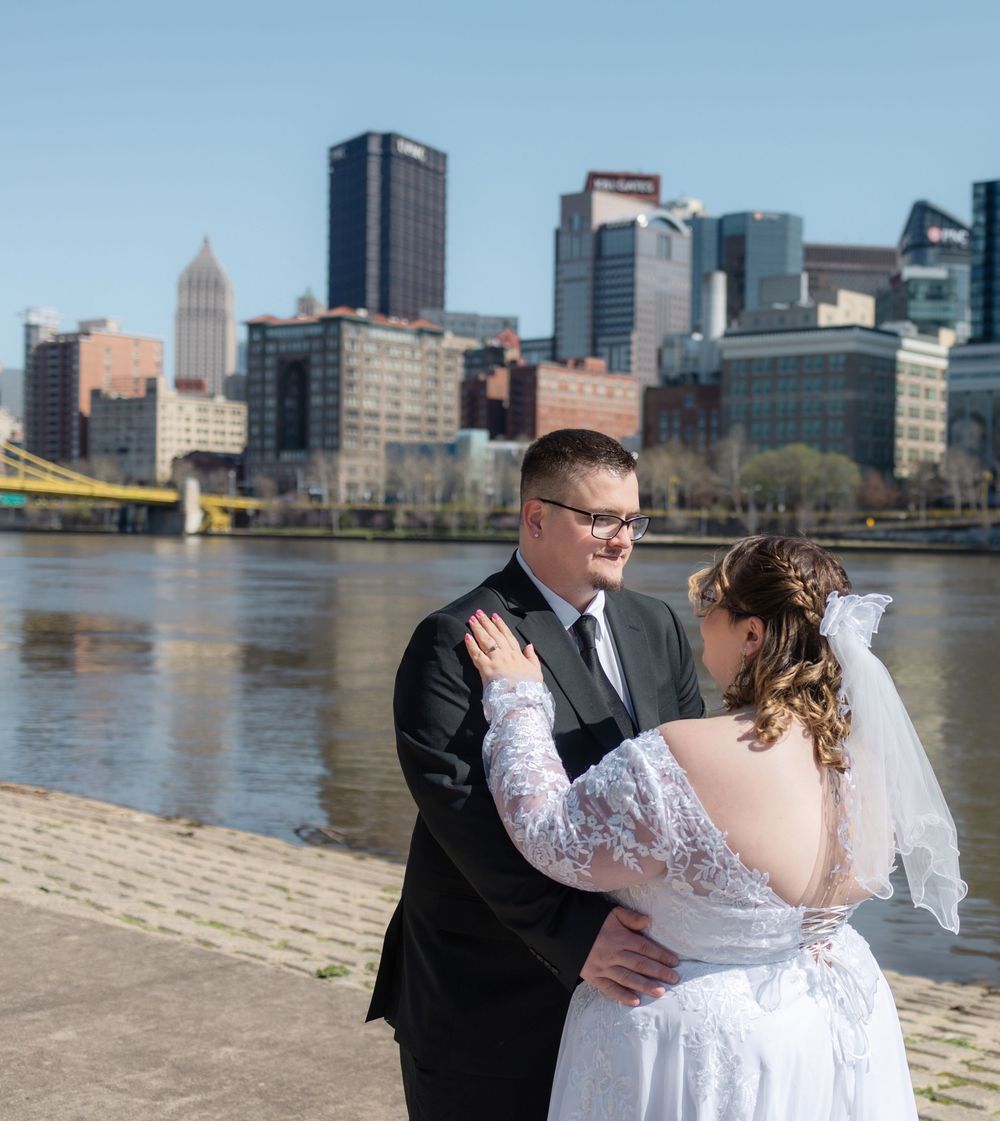 Bride and groom embrace on riverfront with Pittsburgh skyline backdrop on sunny wedding day.