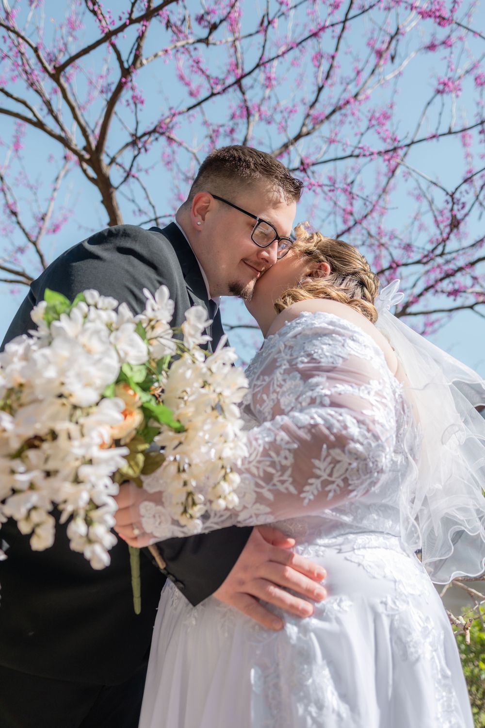 Bride and groom share a romantic kiss under a blooming pink cherry blossom tree, bride holding white floral bouquet.