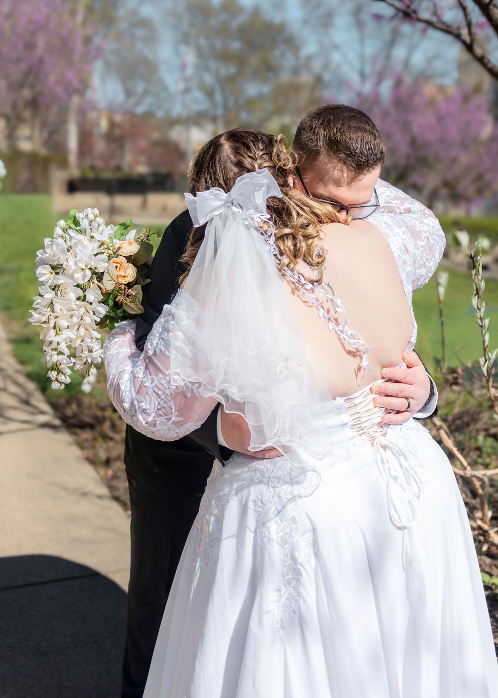 Bride and groom sharing a romantic kiss outdoors, bride wearing lace wedding dress with veil and holding a white bouquet.