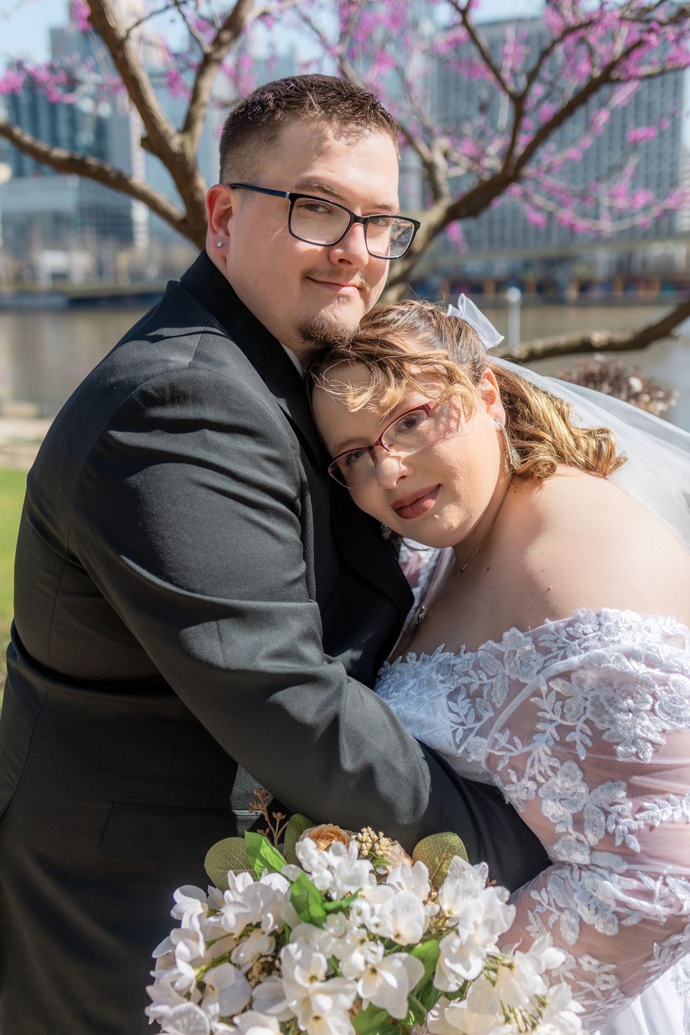 Couple in wedding attire embracing outdoors near blooming tree, bride holding white bouquet, both wearing glasses.