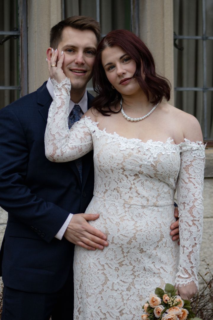 Bride in white lace off-shoulder gown with pearl necklace poses with groom in navy suit at outdoor wedding.