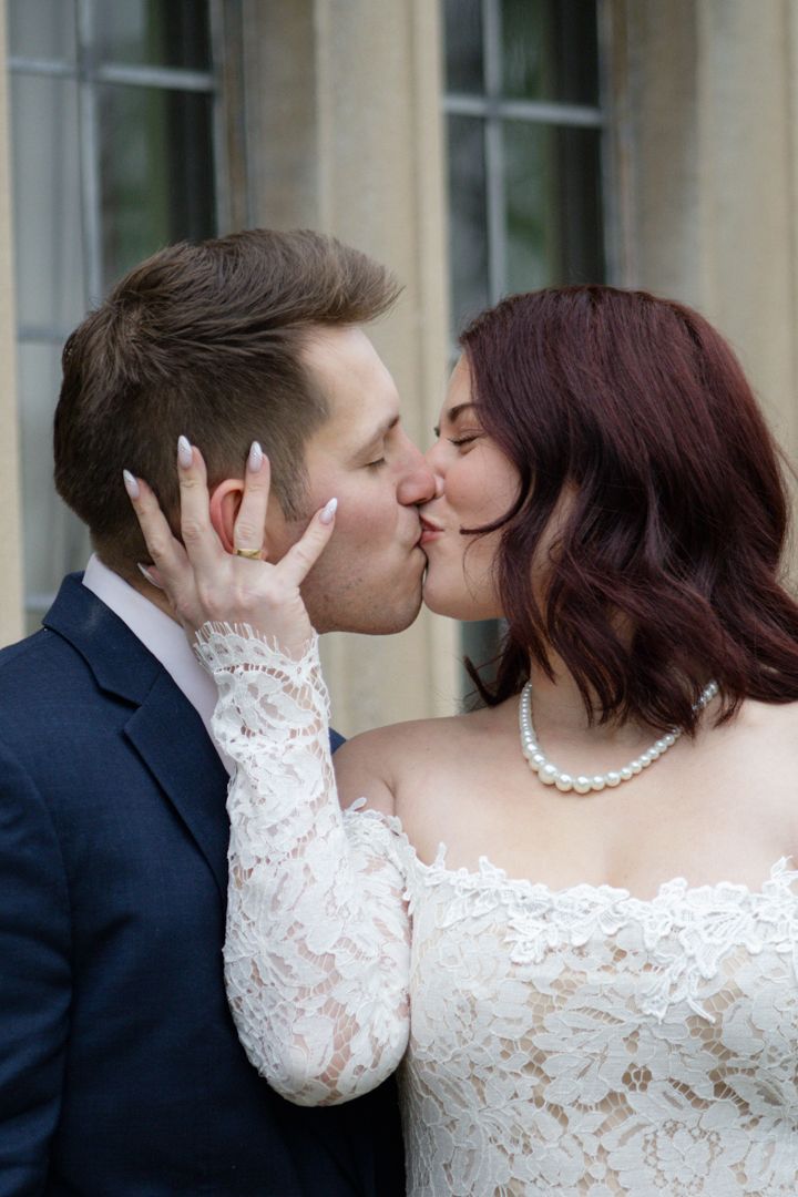 Bride and groom sharing a romantic kiss, bride wearing lace off-shoulder gown and pearl necklace.
