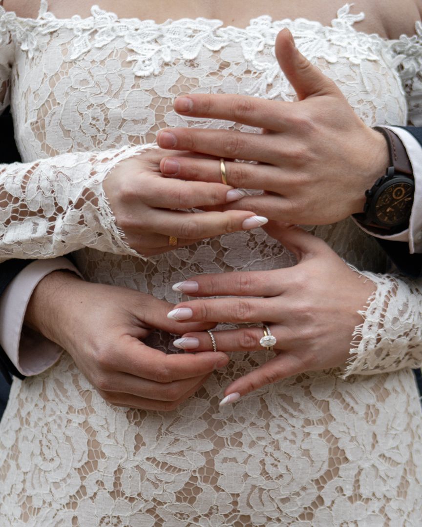 Close-up of bride and groom hands embracing, showing wedding rings and elegant lace wedding dress.