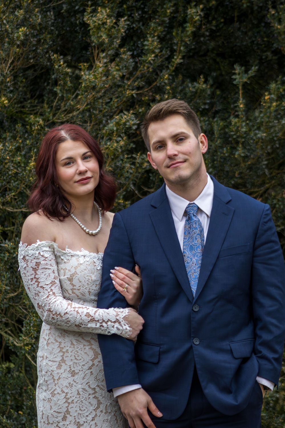 Bride in lace off-shoulder gown and groom in navy suit with blue tie pose together outdoors near green hedges.