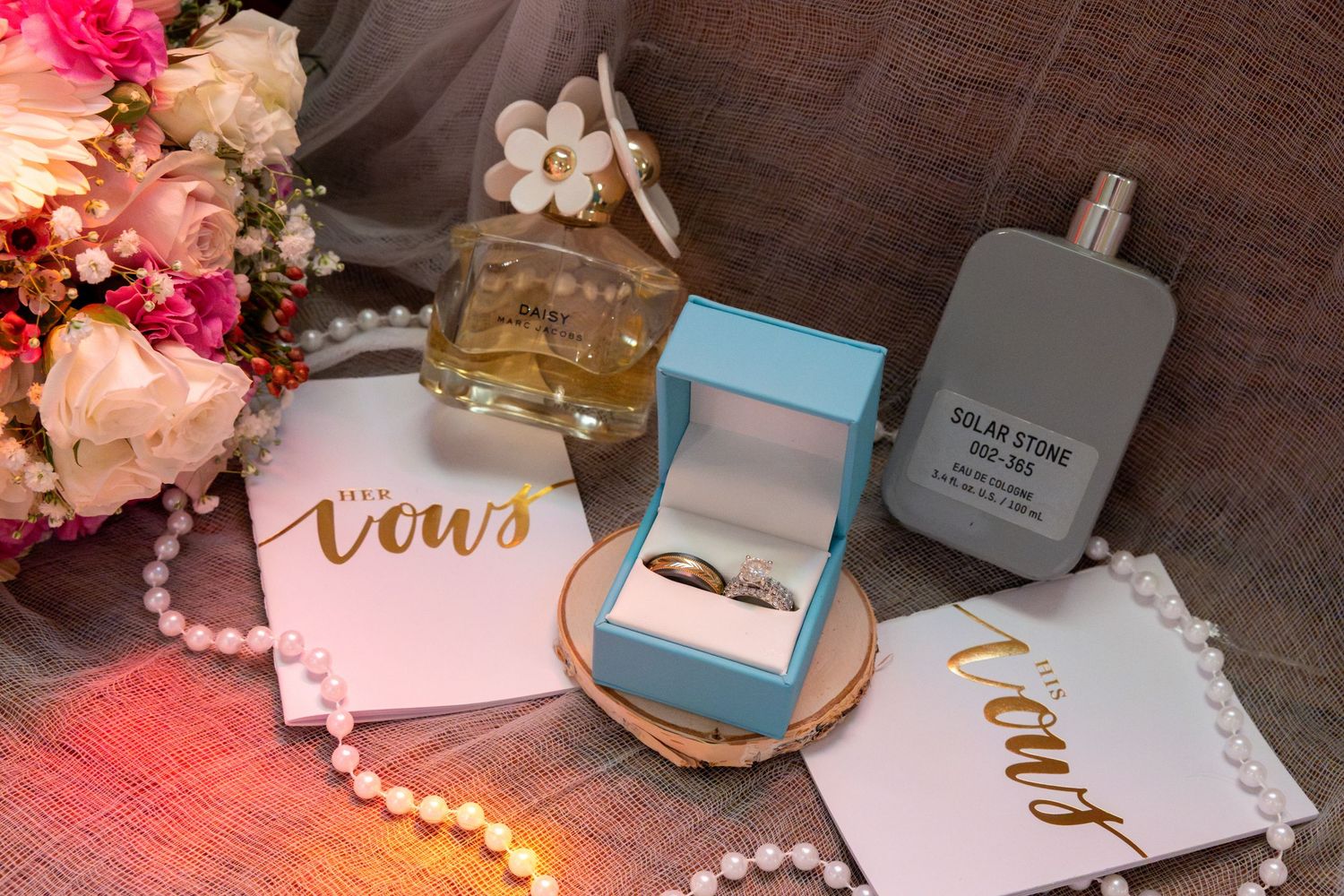 Wedding flat lay with gold rings in blue box, vow books, pearl necklace, perfume bottles, and pink flowers.