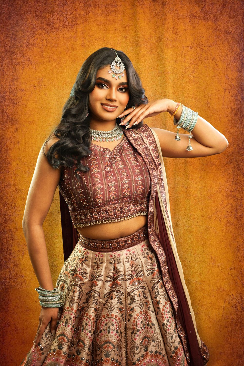 Traditional Indian lehenga outfit in burgundy with intricate embroidery and jewelry styling against warm orange background.