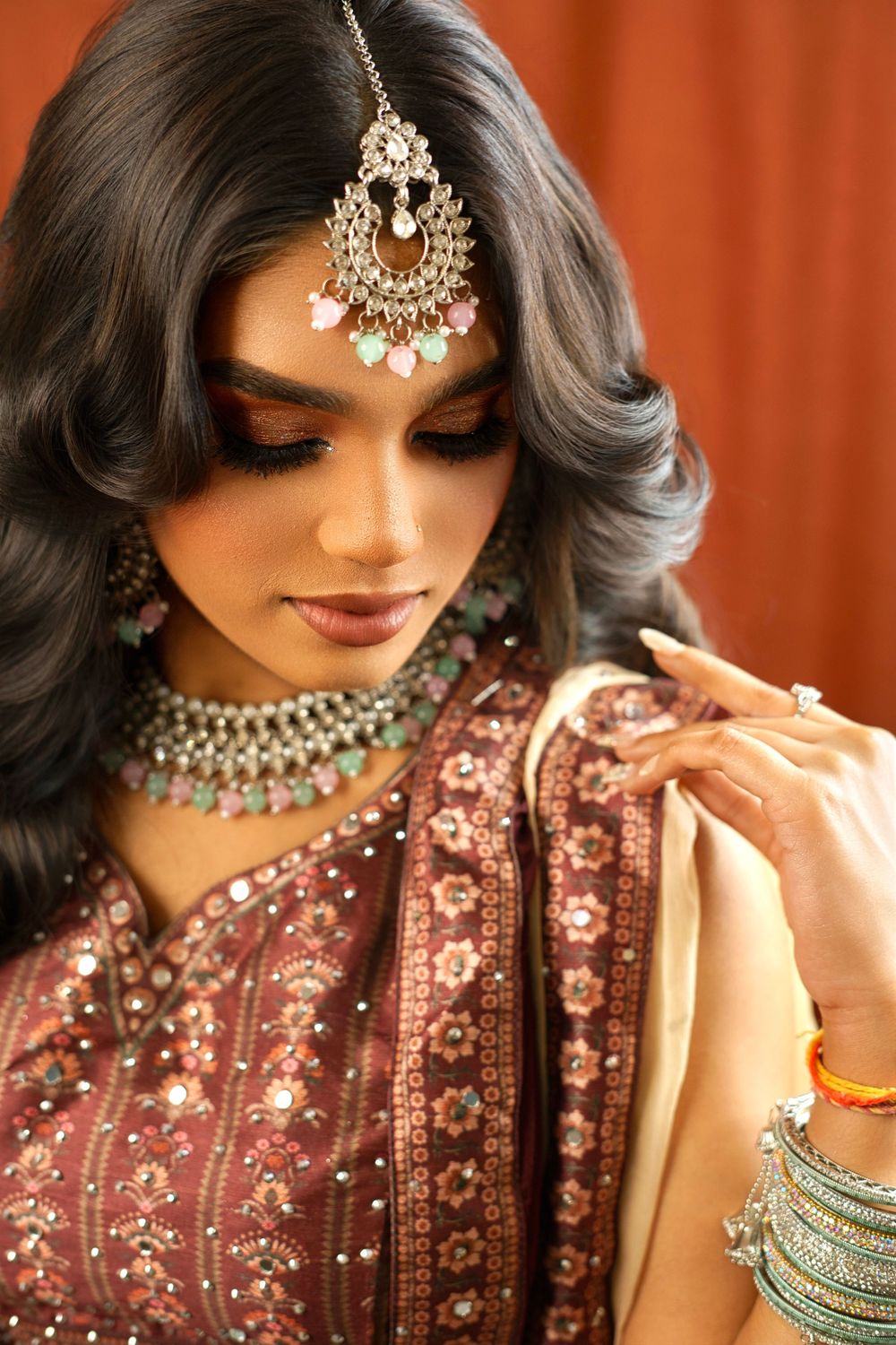 Close-up details of traditional Indian bridal jewelry and embroidered clothing in burgundy against an orange backdrop.