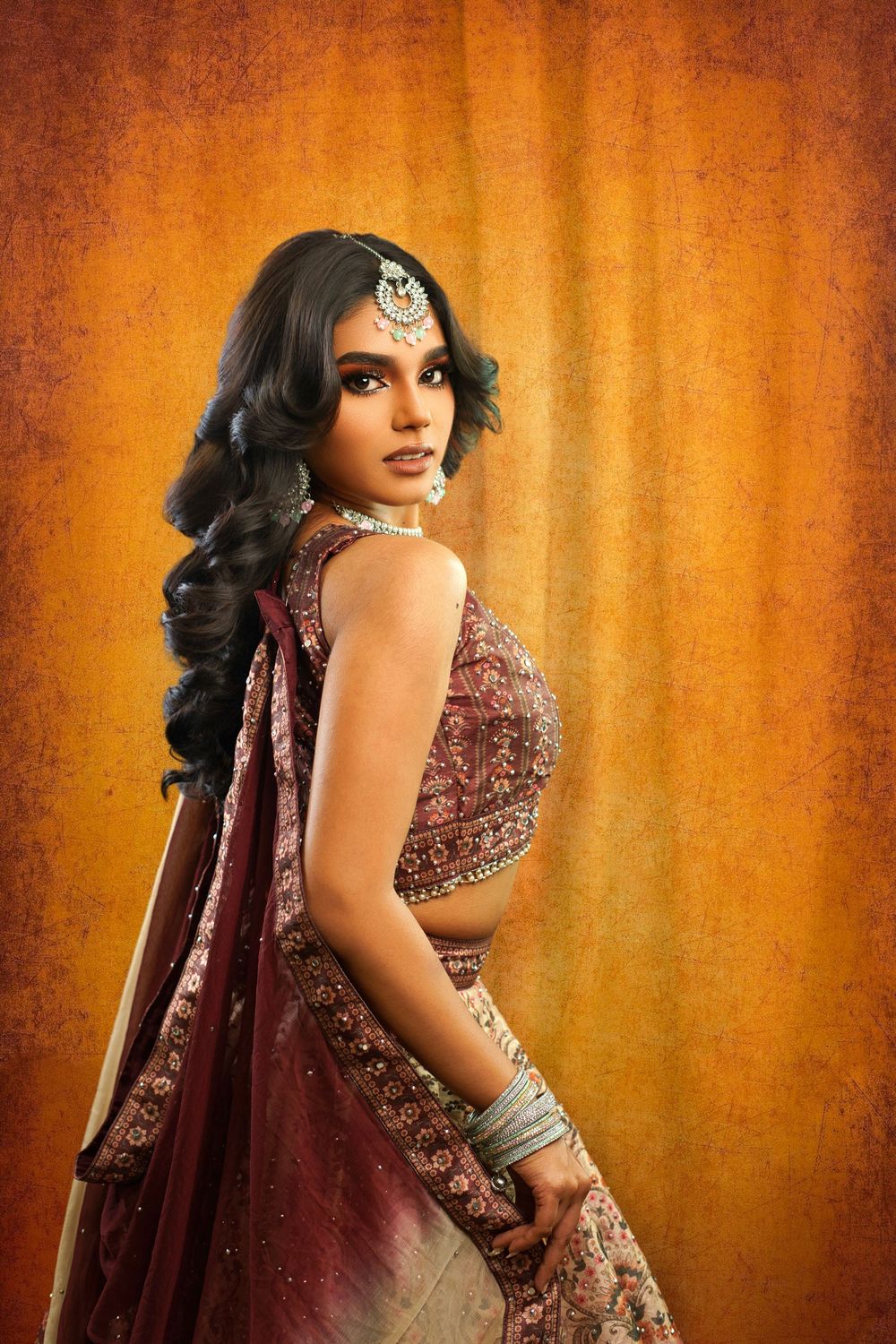 A woman in an elegant burgundy and gold Indian lehenga poses against a warm orange backdrop.