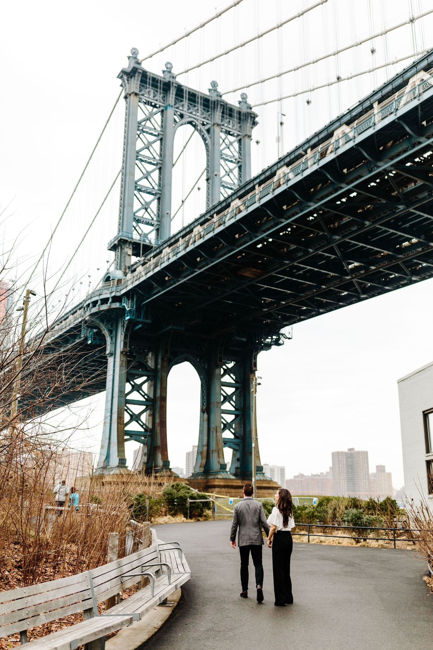 Couple walking along a path beneath the Manhattan Bridge on an overcast day.