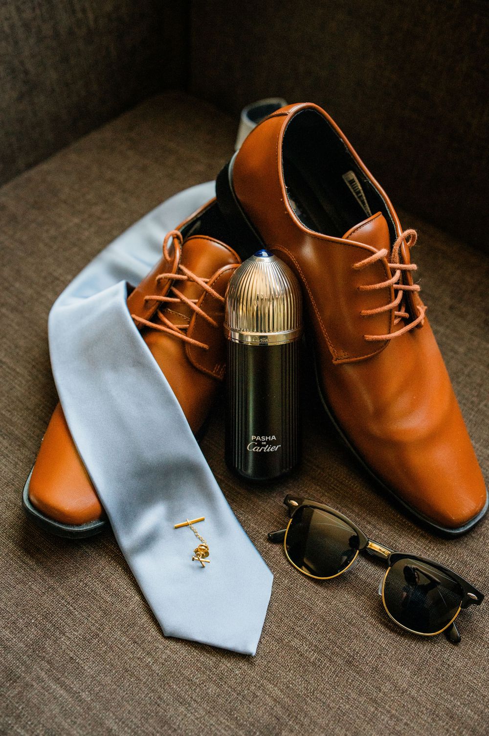 Brown leather dress shoes with a light blue tie and cologne bottle arranged on textured fabric.