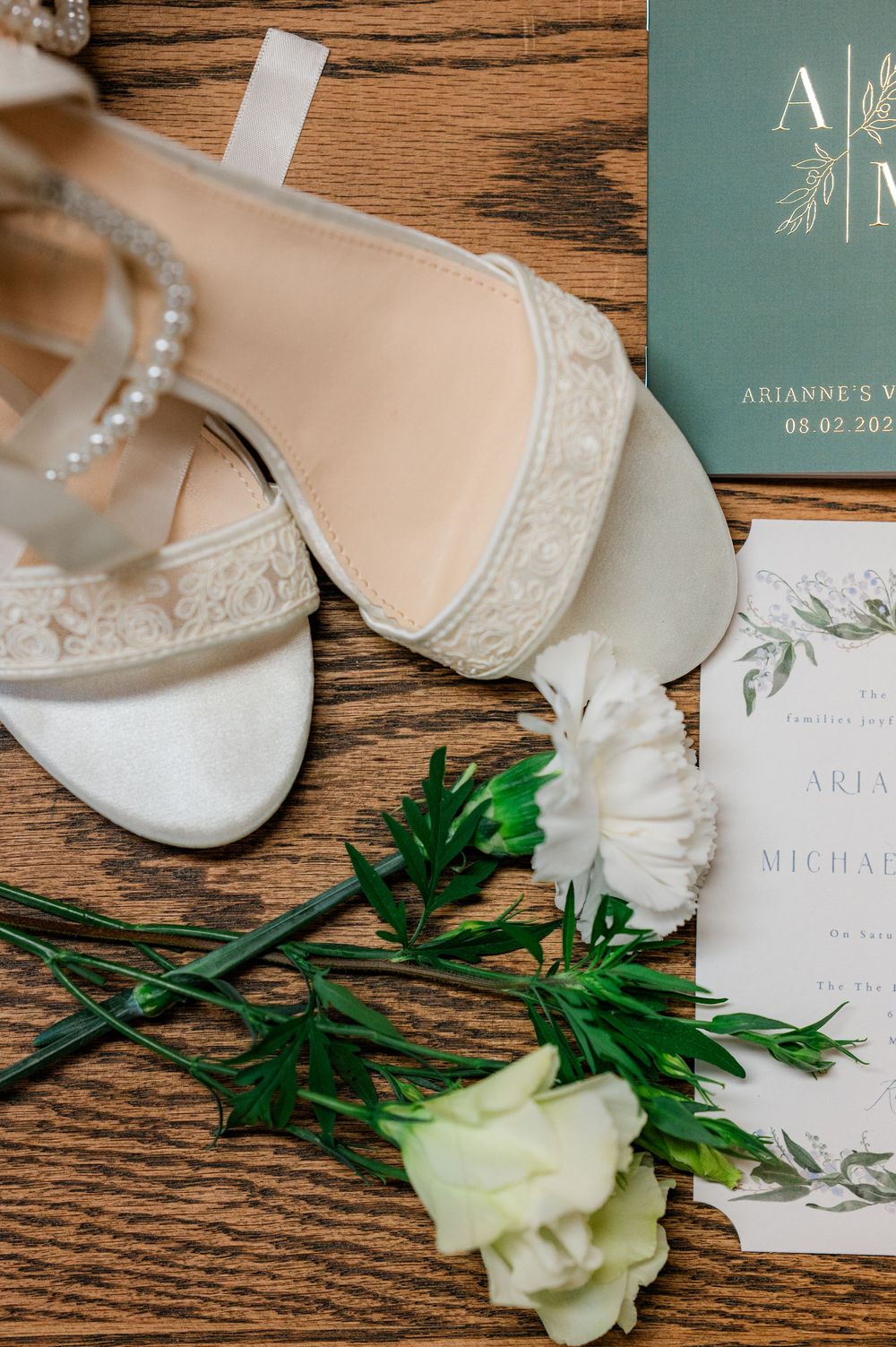 Wedding invitation and bridal accessories including white shoes and roses arranged on wooden surface.