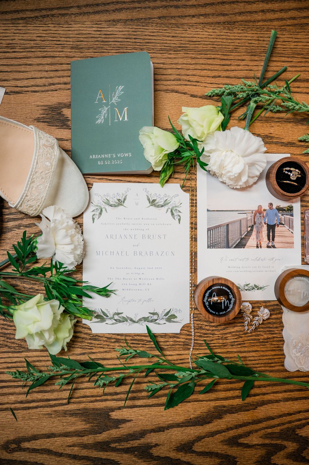 Elegant wedding invitation suite with white flowers and greenery laid out on rustic wooden surface.
