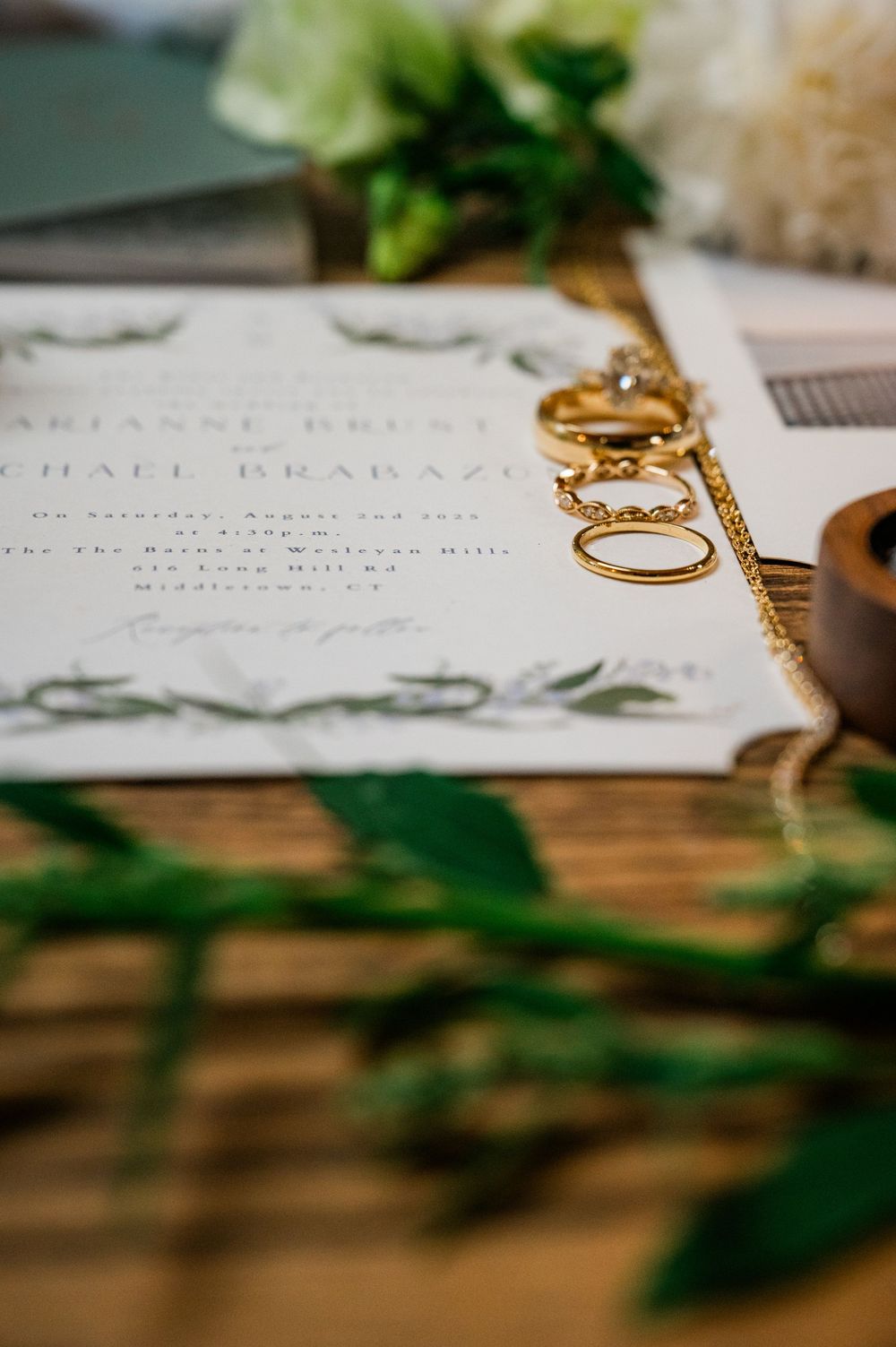 Wedding invitation with gold rings and greenery details on wooden surface.