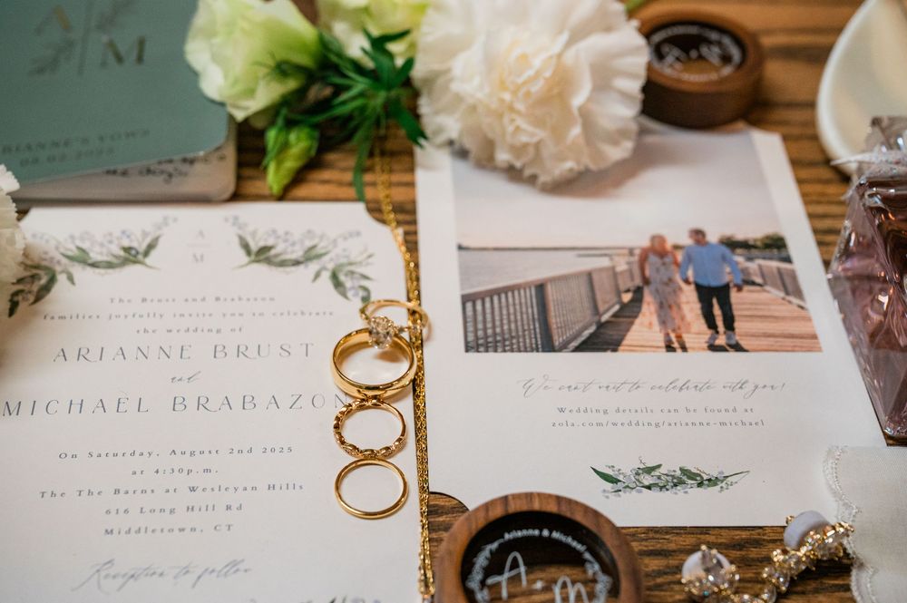 Wedding invitation suite with gold rings and greenery on a wooden surface.