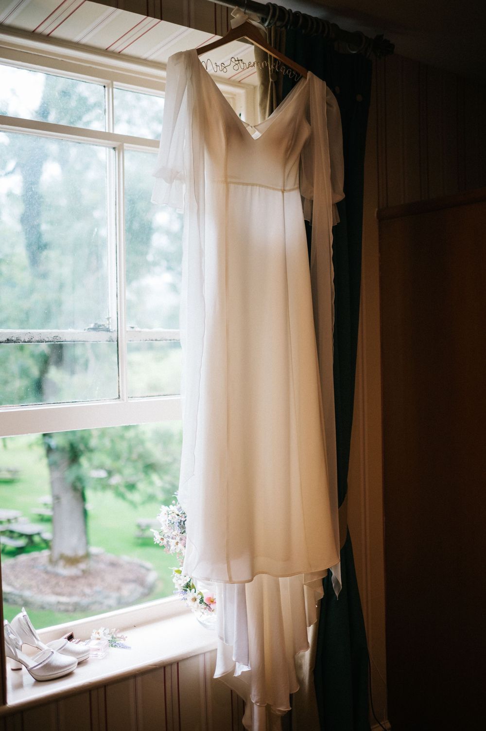 An elegant white wedding dress hangs by a window in natural light.