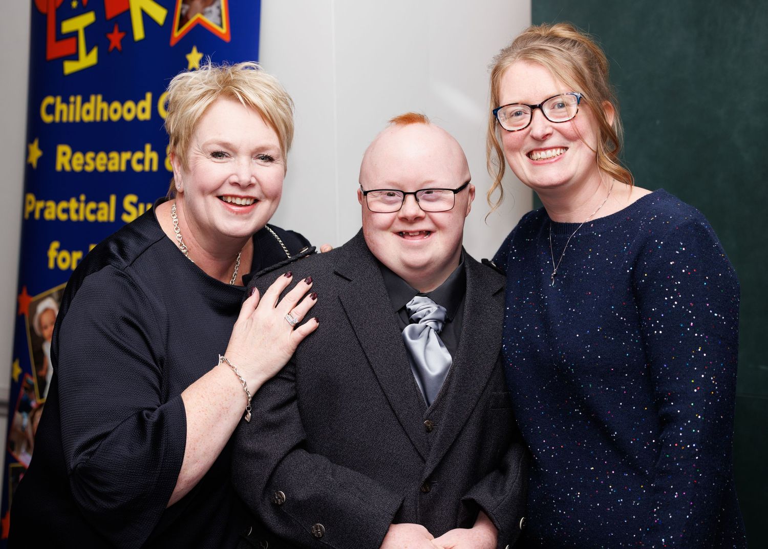 Three people pose together at an event with a blue banner visible in the background.