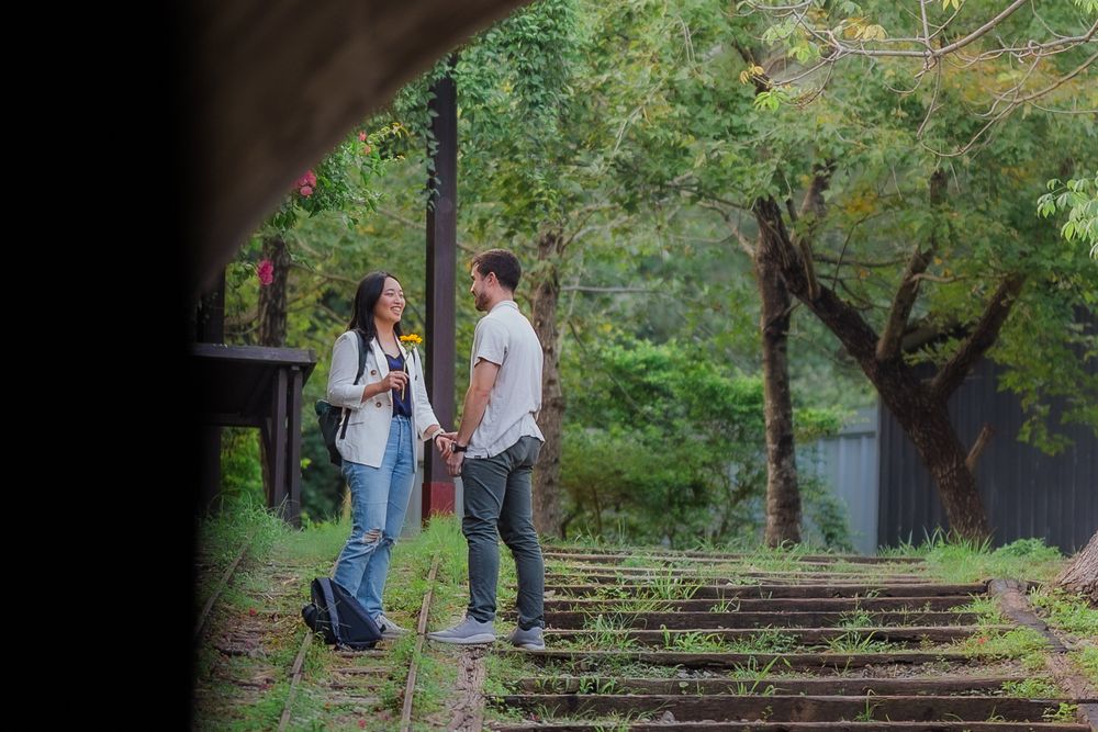 A couple stands together in a lush green garden setting during what appears to be an engagement photoshoot.