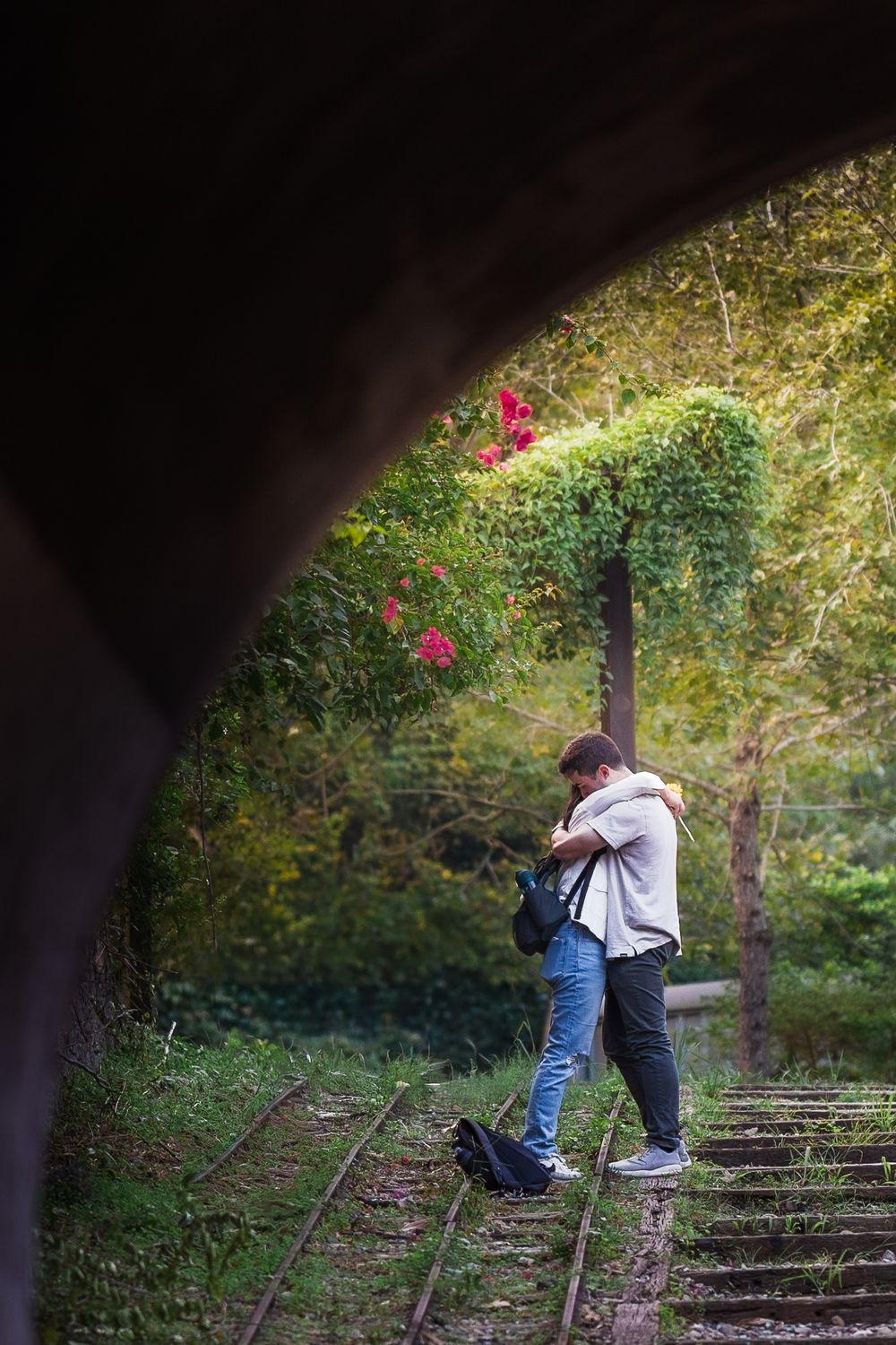 A couple embraces on old train tracks surrounded by lush greenery and pink flowers in a romantic forest setting.