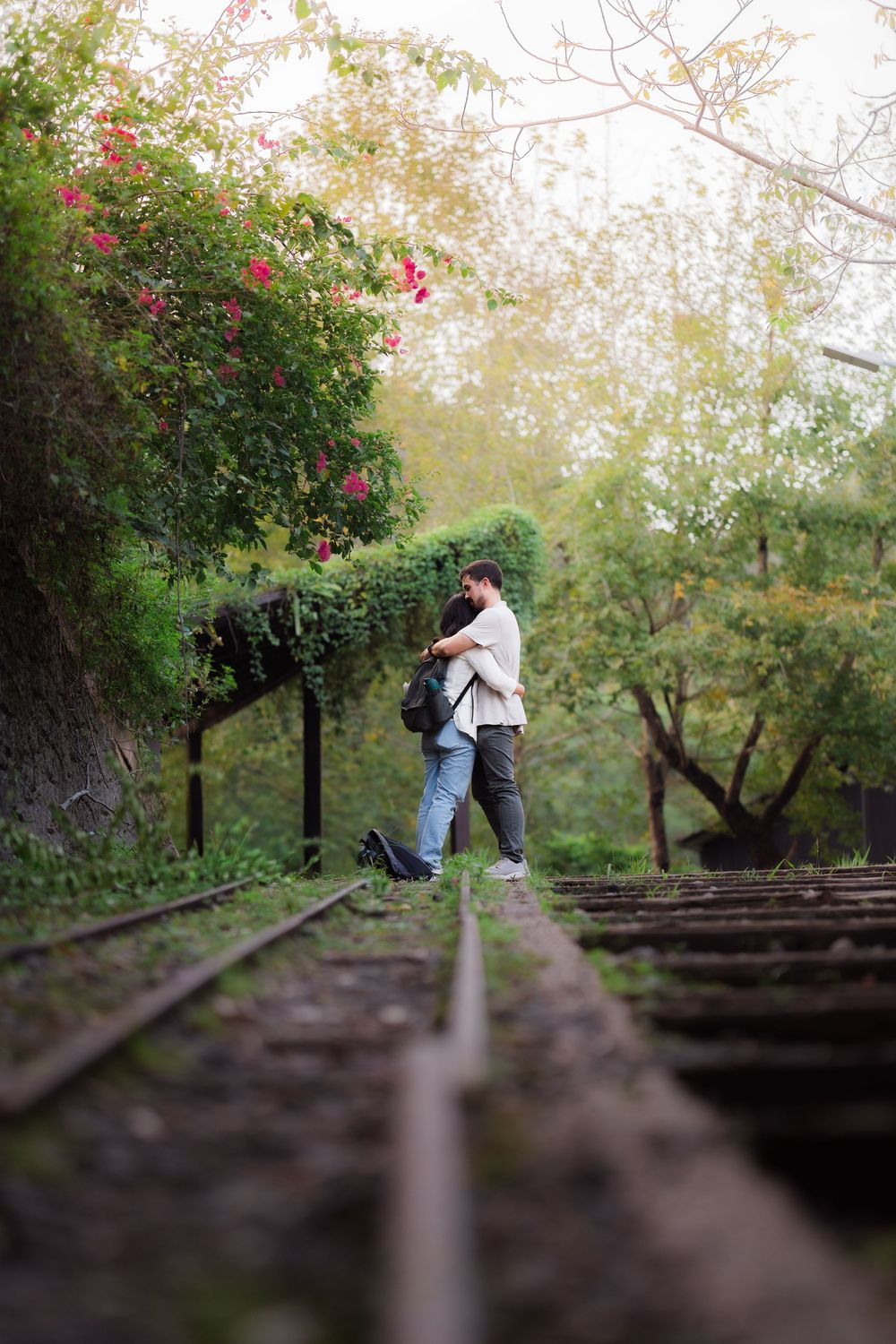 Couple sits on wooden railroad tracks surrounded by lush greenery and flowering trees in a romantic setting.