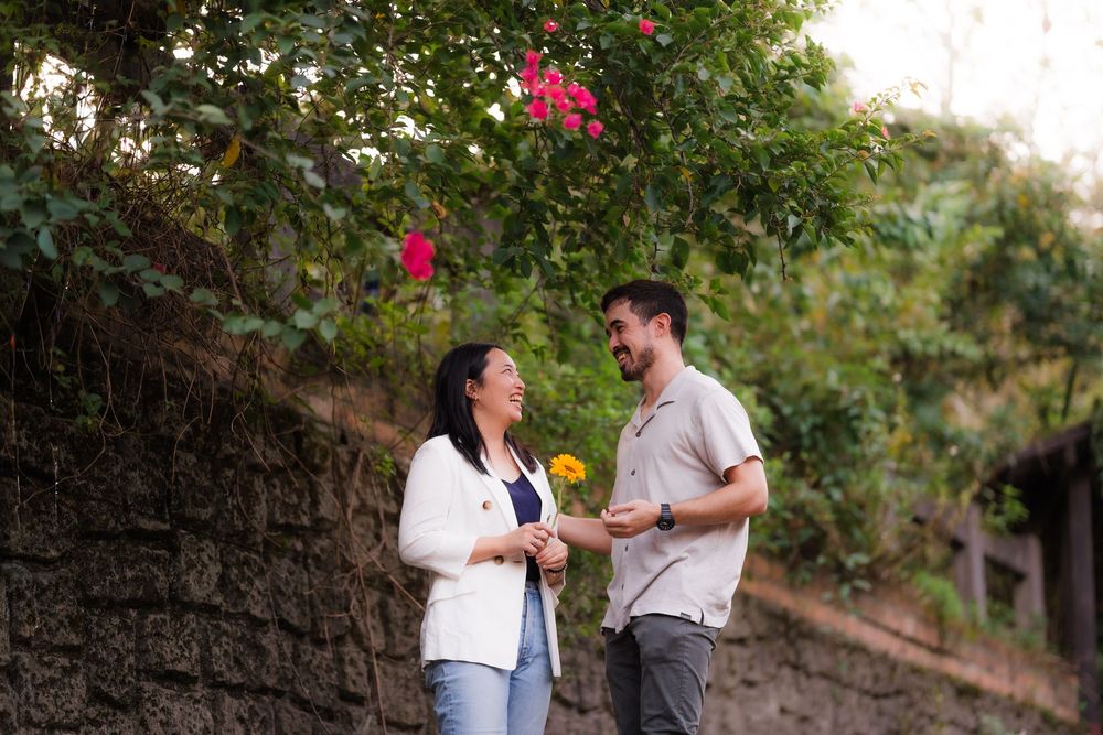Couple shares a sweet moment exchanging flowers under blooming trees in a garden setting.
