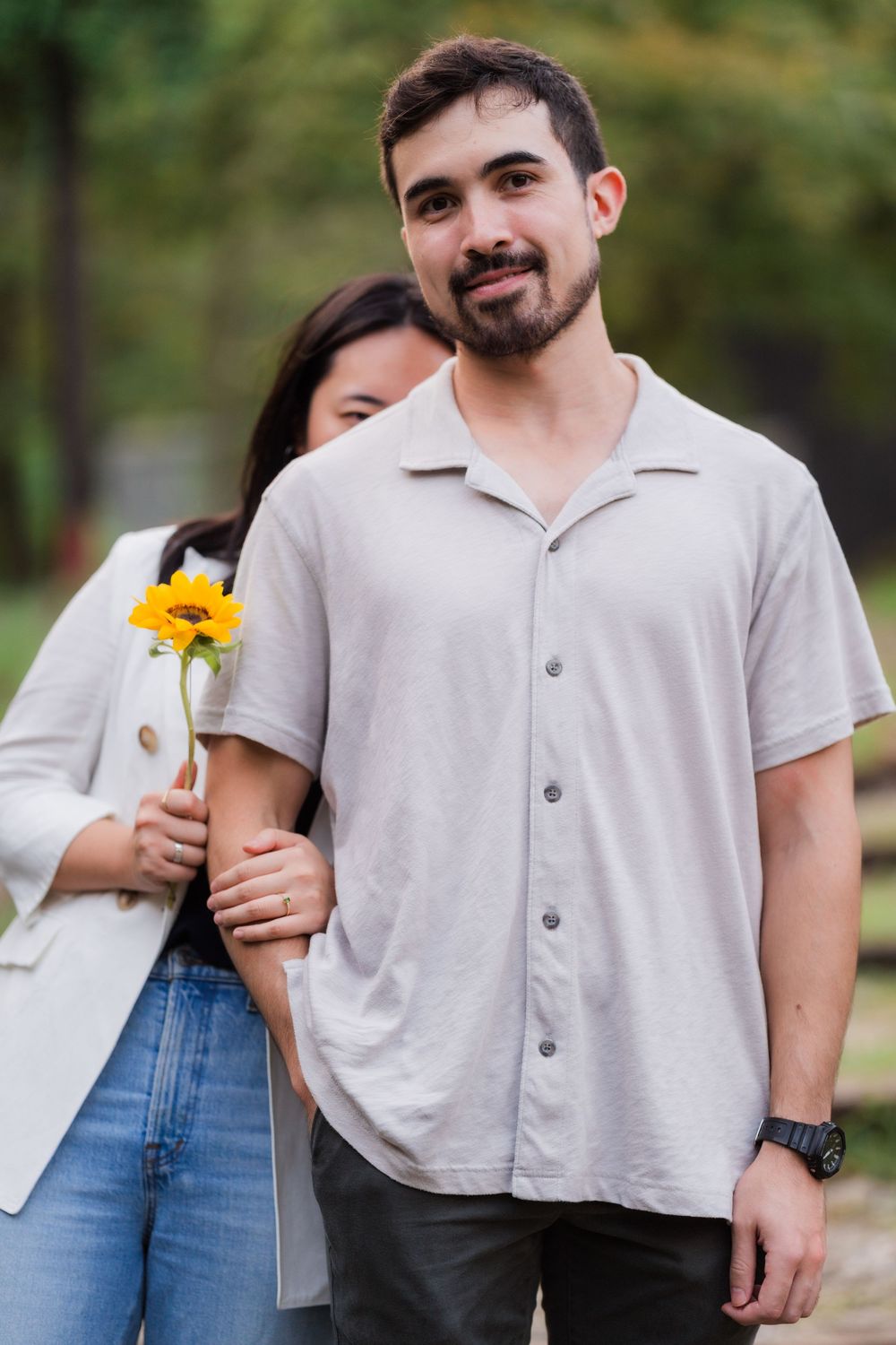 A couple in casual attire shares an intimate moment outdoors with a bright yellow sunflower.