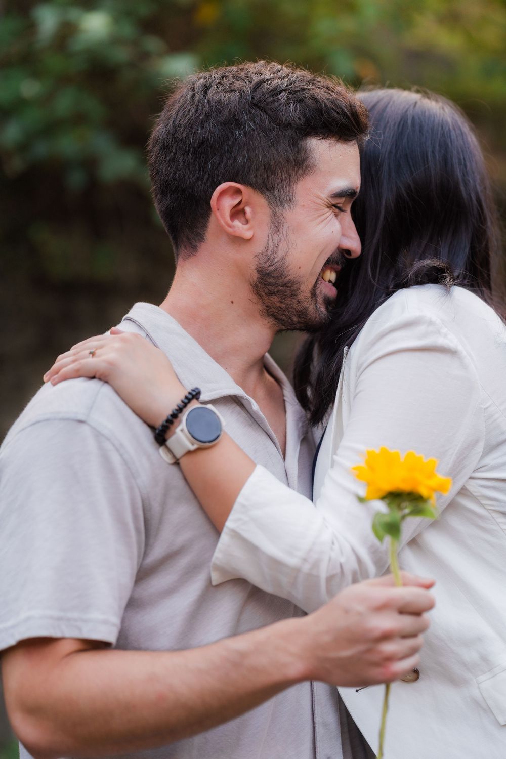 Romantic couple in white shirts embracing while holding a yellow sunflower in an outdoor setting.