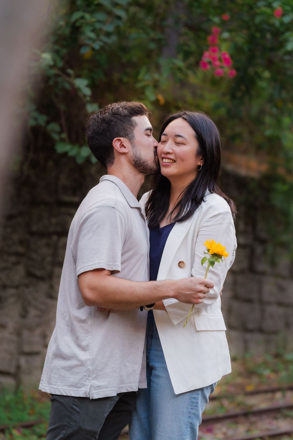 A couple shares an intimate moment outdoors while holding a yellow flower, surrounded by greenery and stone walls.