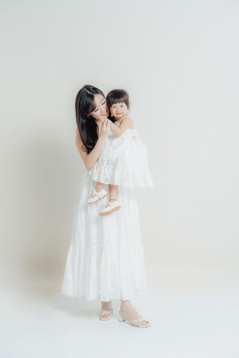Two figures in matching white dresses embrace each other against a light background in a minimalist studio setting.
