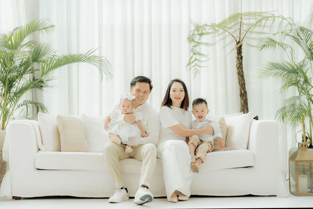 A family sits together on a white couch surrounded by palm plants in a bright, minimalist interior setting.