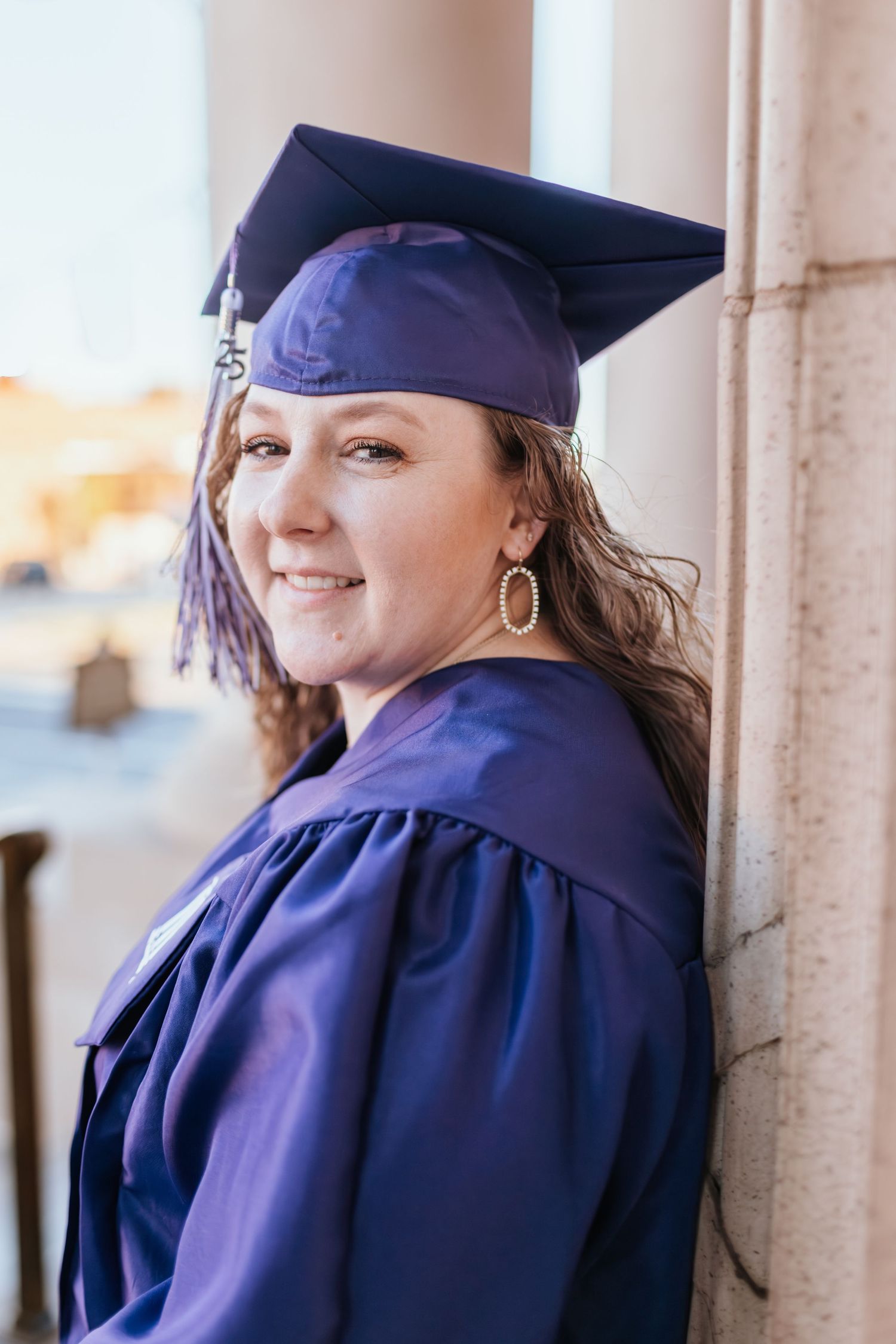 A graduate in navy blue cap and gown poses for photos against stone columns in natural lighting.