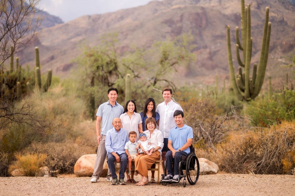 Multi-generational family portrait in Arizona desert with saguaro cacti, including a young man in a wheelchair, smiling outdoors.