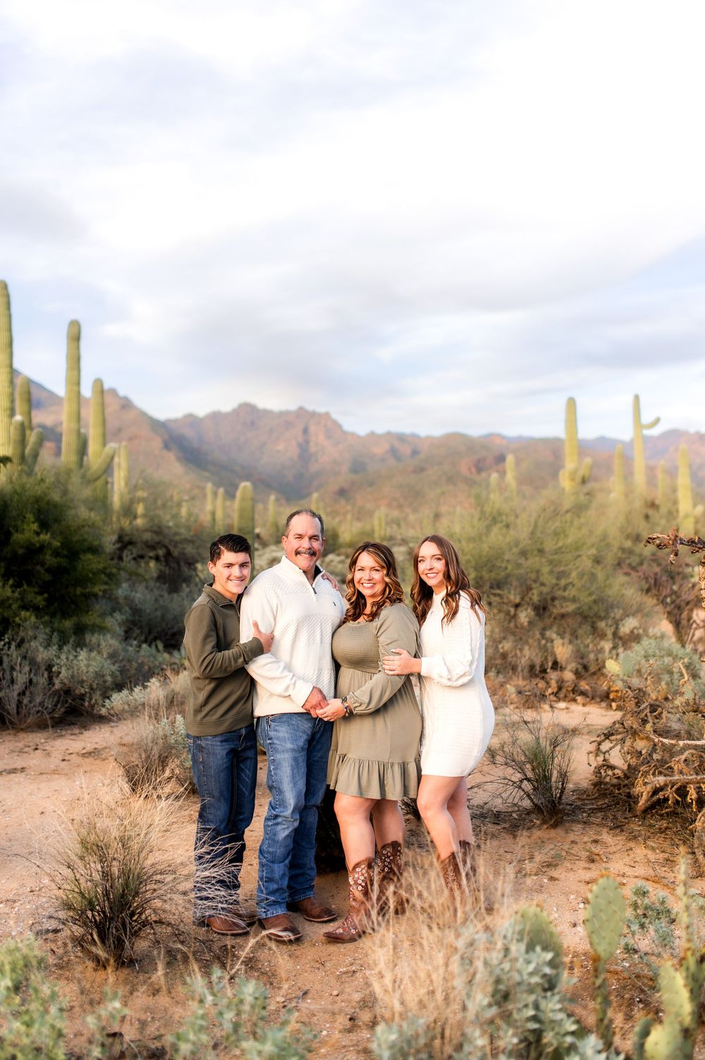 Family of four smiles together in Arizona desert landscape with saguaro cacti and mountain backdrop.
