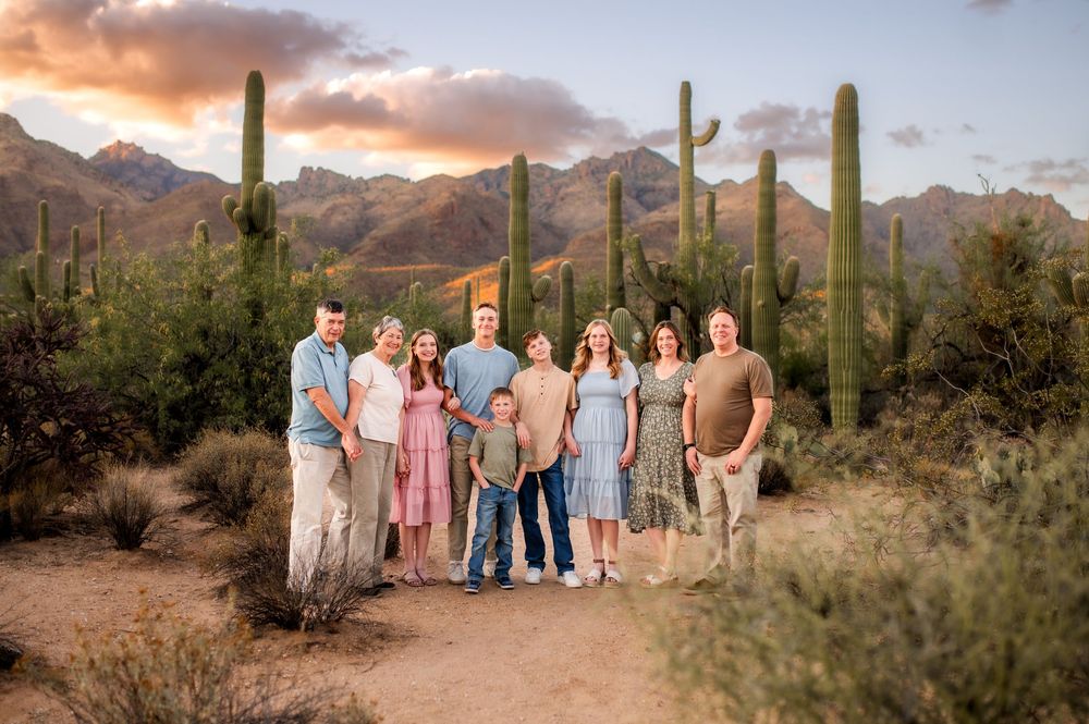 Extended family of eight poses among tall saguaro cacti with colorful sunset mountains in Arizona desert.
