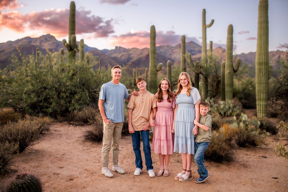 Five siblings smile together in Arizona desert landscape with towering saguaro cacti and pink sunset sky.