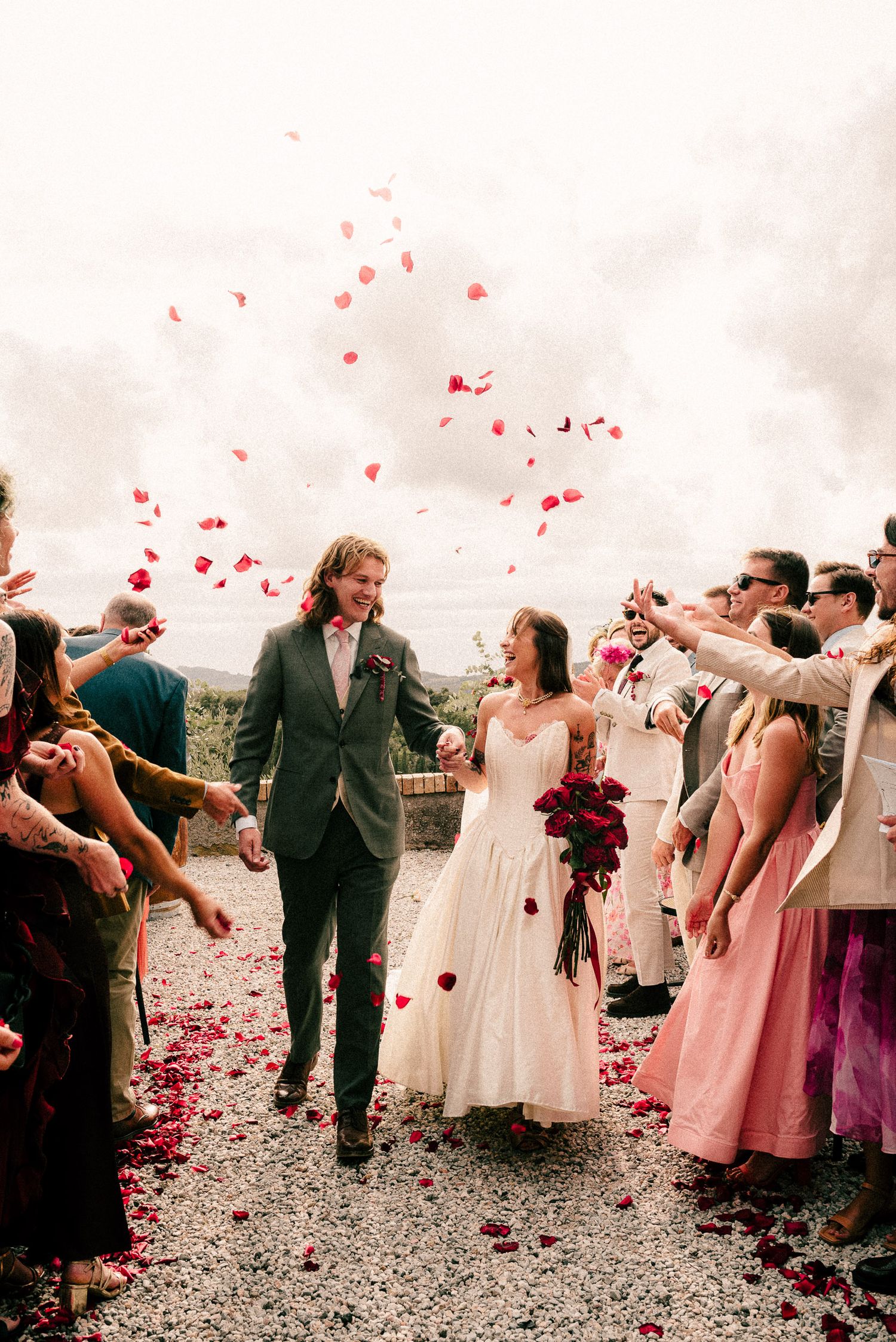 Wedding party celebrates on a beach as guests shower them with red rose petals in a candid sequence of photos.