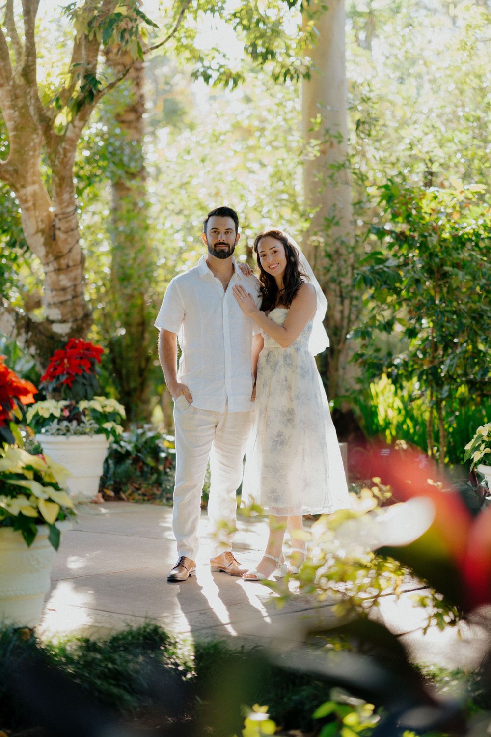 A couple in white attire walks along a sunlit garden path surrounded by lush greenery and red flowers.