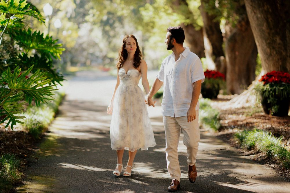 A couple strolls hand in hand along a garden path lined with lush greenery and Spanish moss covered trees.