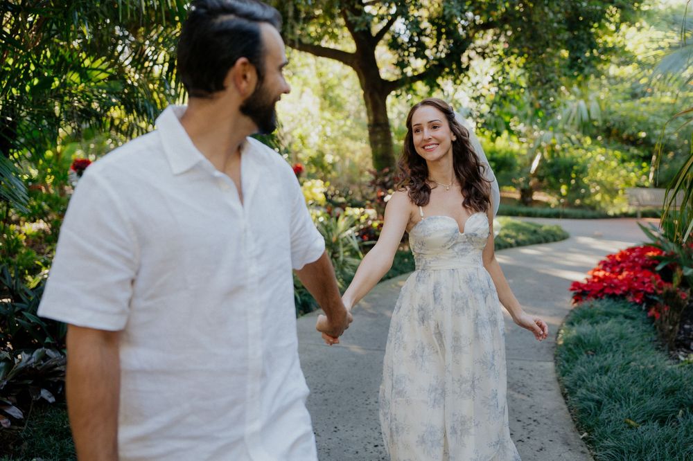 A couple in white outfits poses together in a garden setting during a romantic outdoor photoshoot.