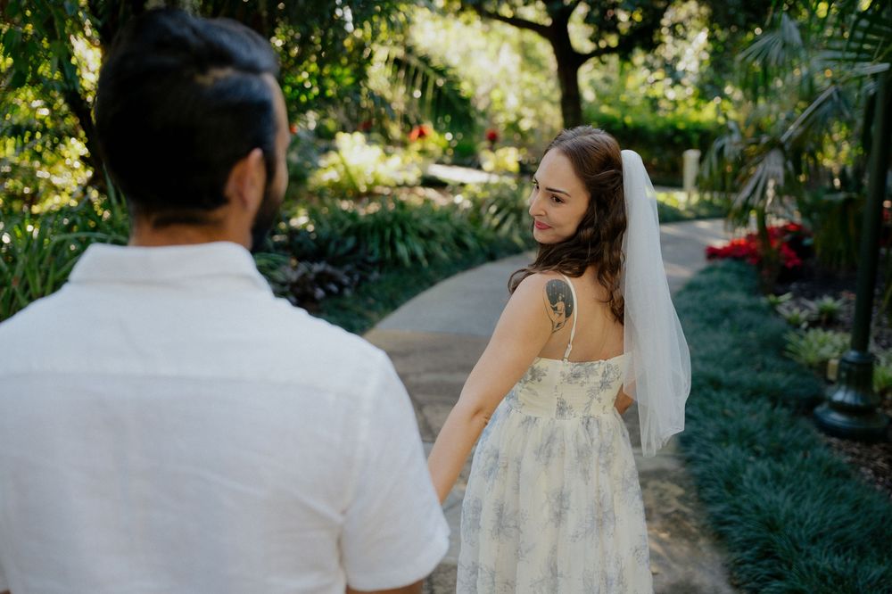 A bride in a floral lace dress looks back while walking along a garden pathway on her wedding day.