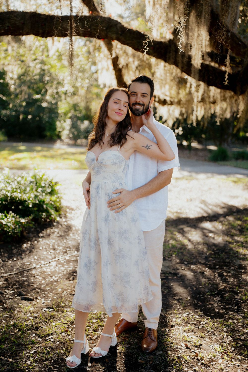 Couple in white outfits embraces in a sun-dappled forest setting during a romantic outdoor photoshoot.