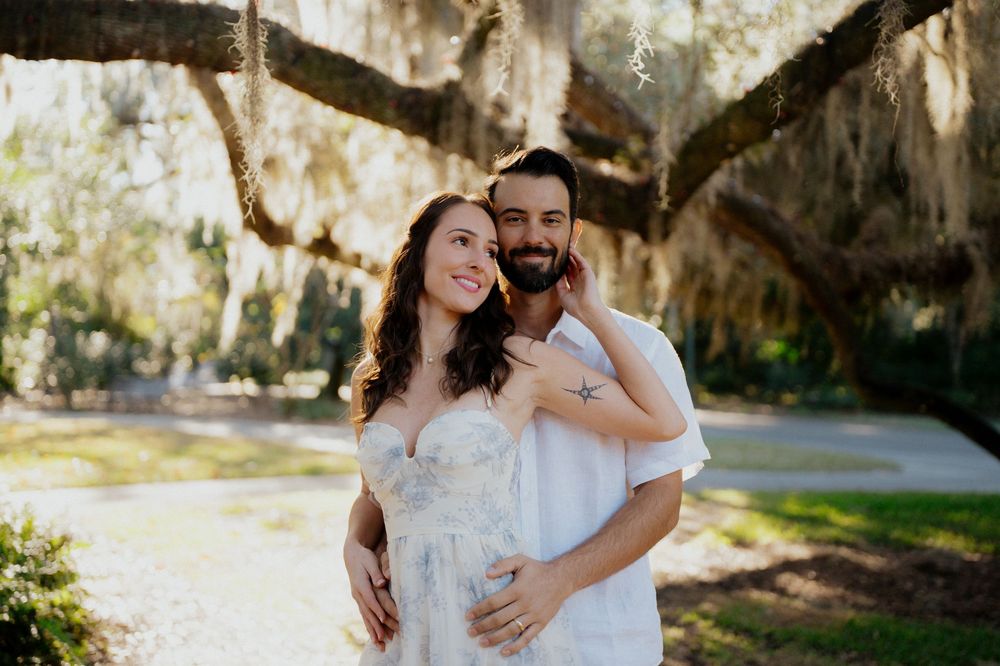 A romantic couple shares a tender moment under Spanish moss draped trees in a sunlit Southern garden setting.