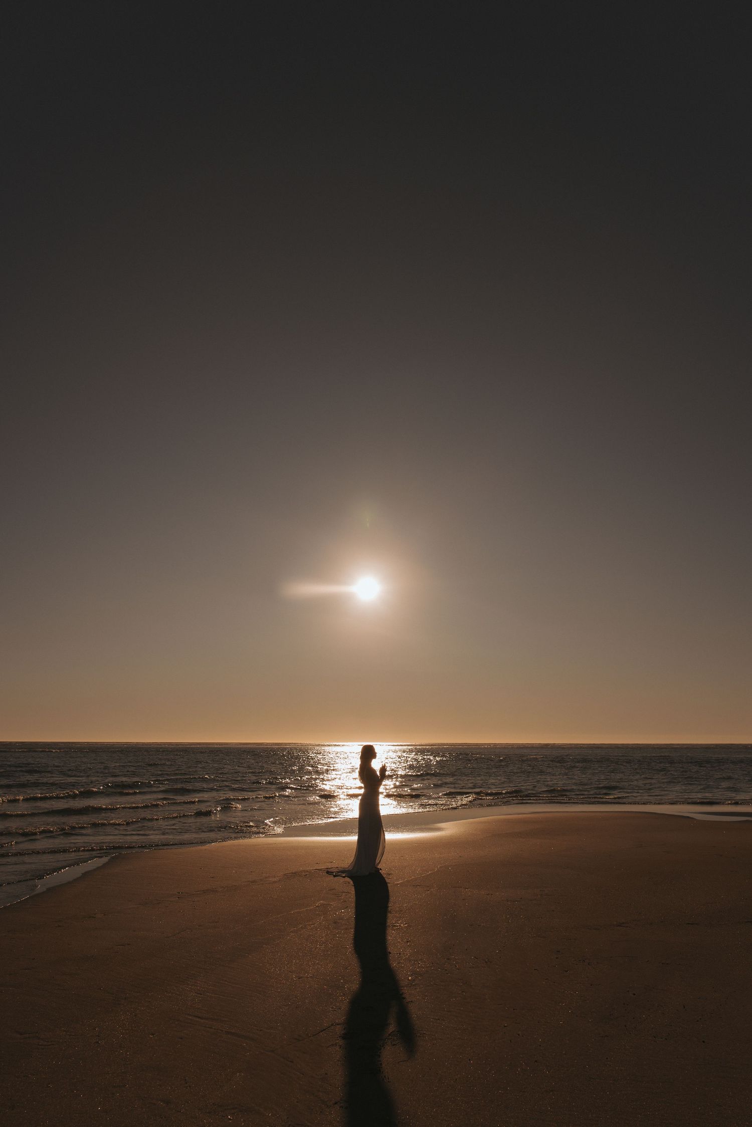 Silhouette of woman in white dress standing on sandy beach with sun reflecting on ocean in New Zealand.