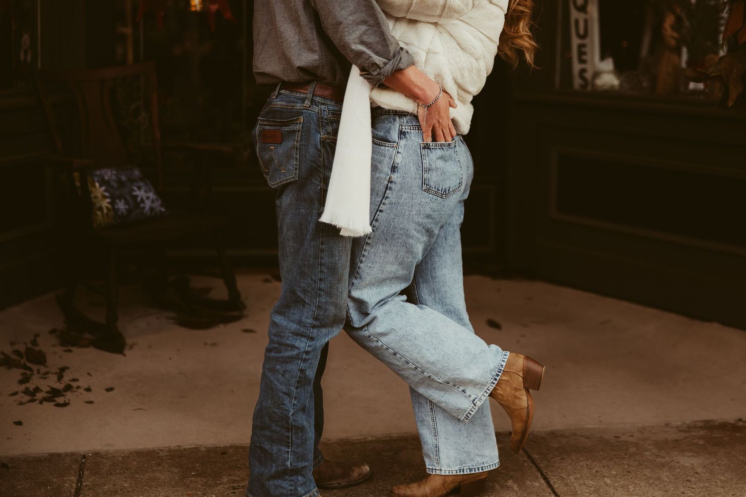Romantic couple in jeans and cowboy boots embracing outside a rustic storefront at dusk.