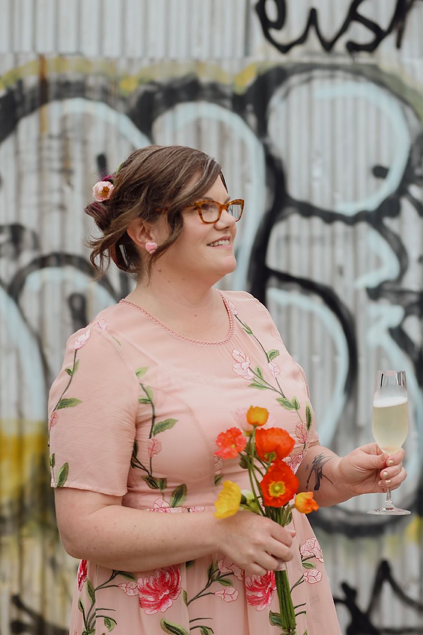 A person in a floral pink dress holds a champagne glass and flowers against a graffiti wall background.