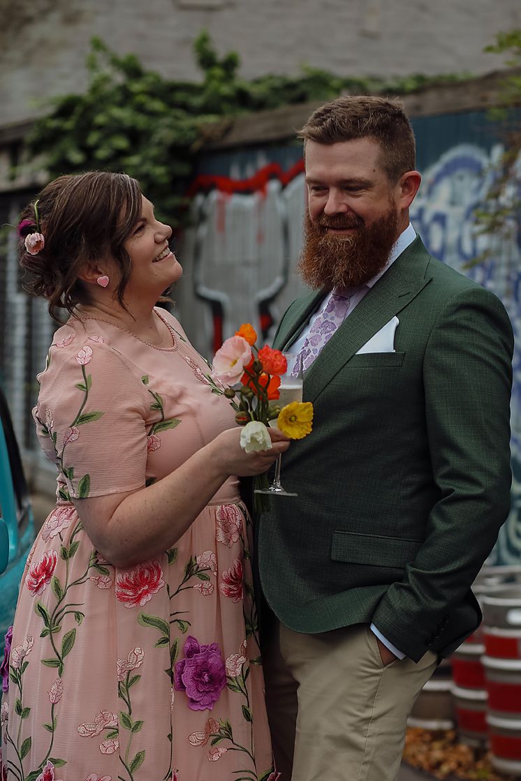 A couple in formal attire shares a joyful moment at an outdoor venue with graffiti walls in the background.