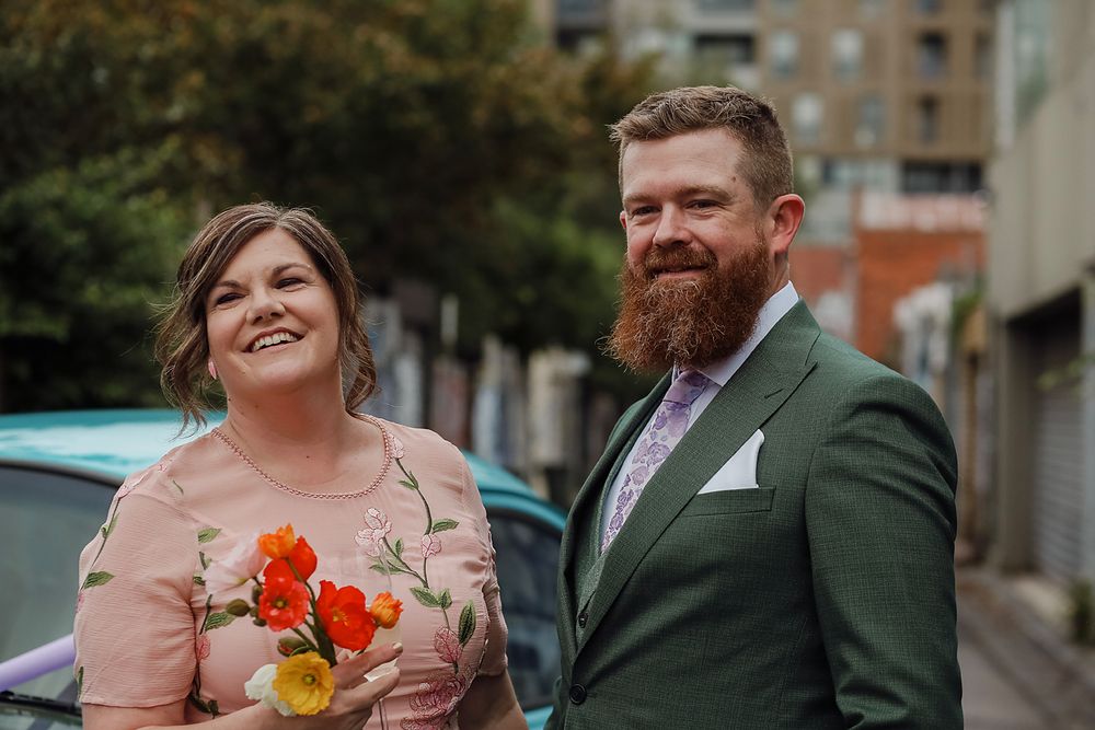 A wedding celebration scene with people in formal attire including a green suit and pink dress against an urban backdrop.