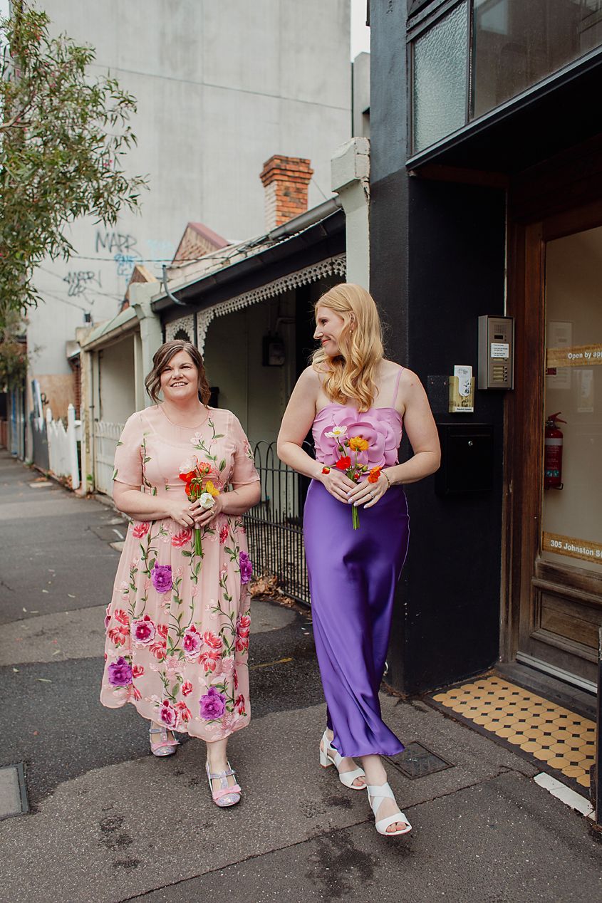 Two people in colorful dresses sit and stand outside a storefront on a city street.