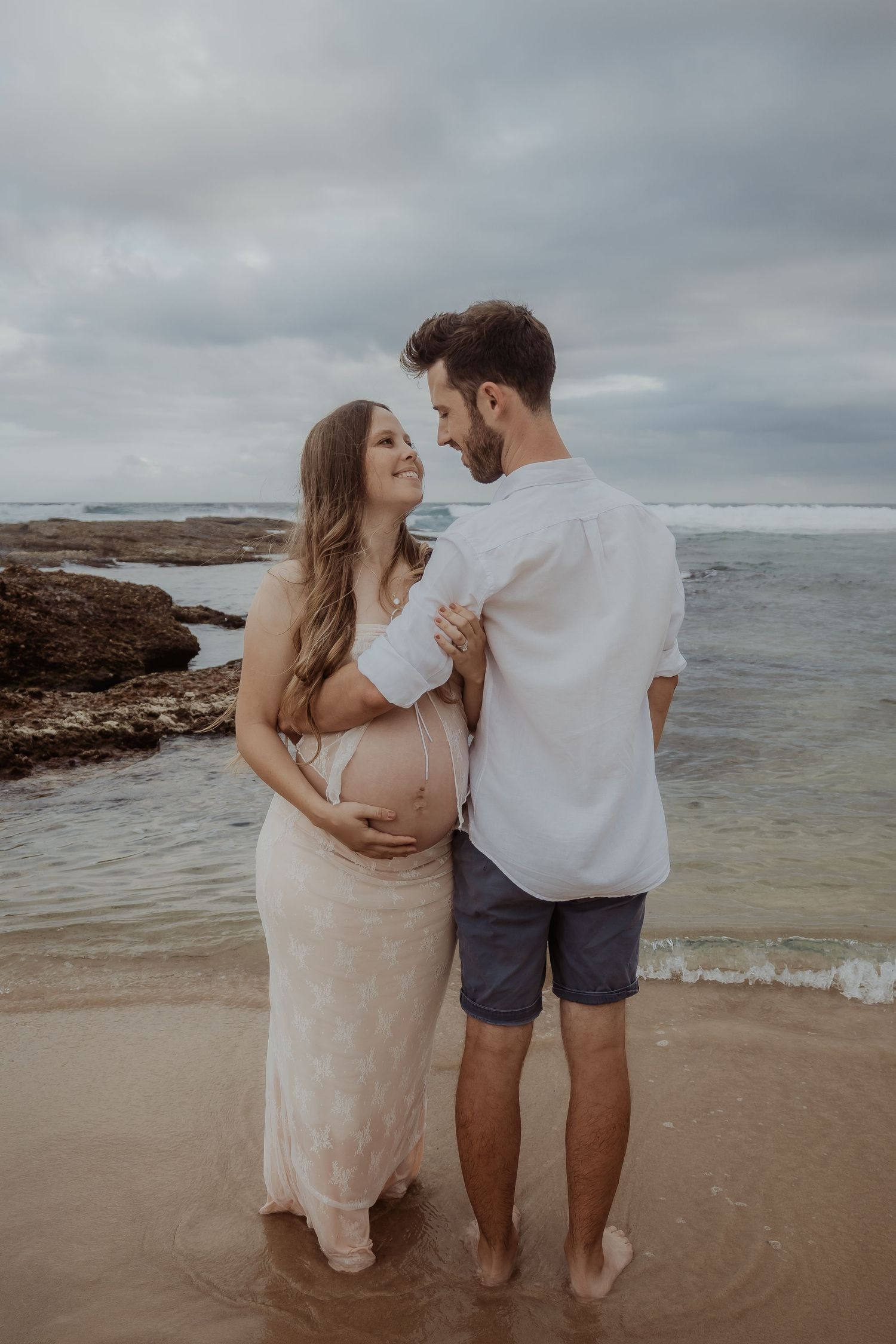 Romantic central coast maternity photoshoot on beach with couple standing in shallow ocean water at sunset.