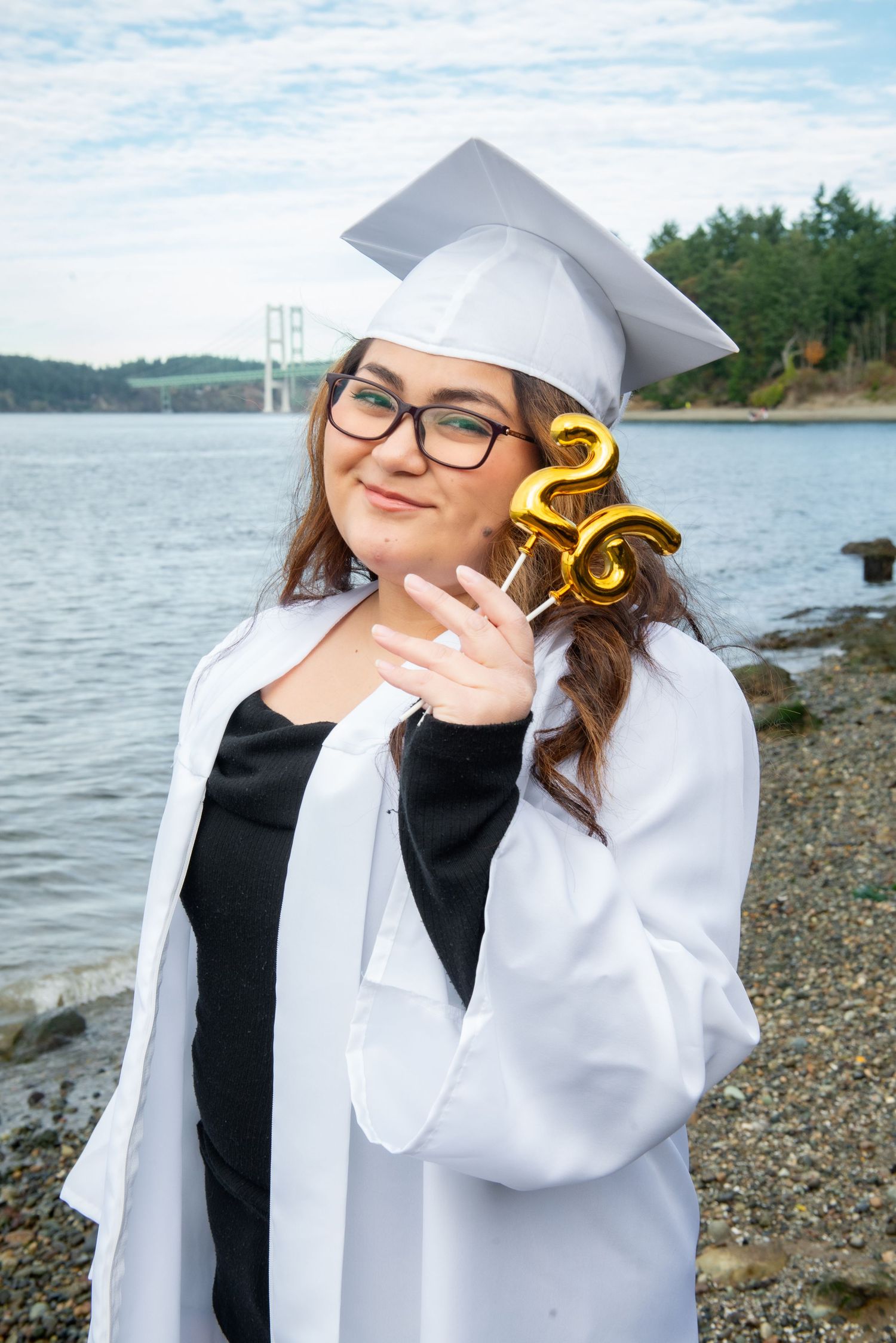 Graduate in white cap and gown poses with decorative numbers on rocky beach with water and trees in background.