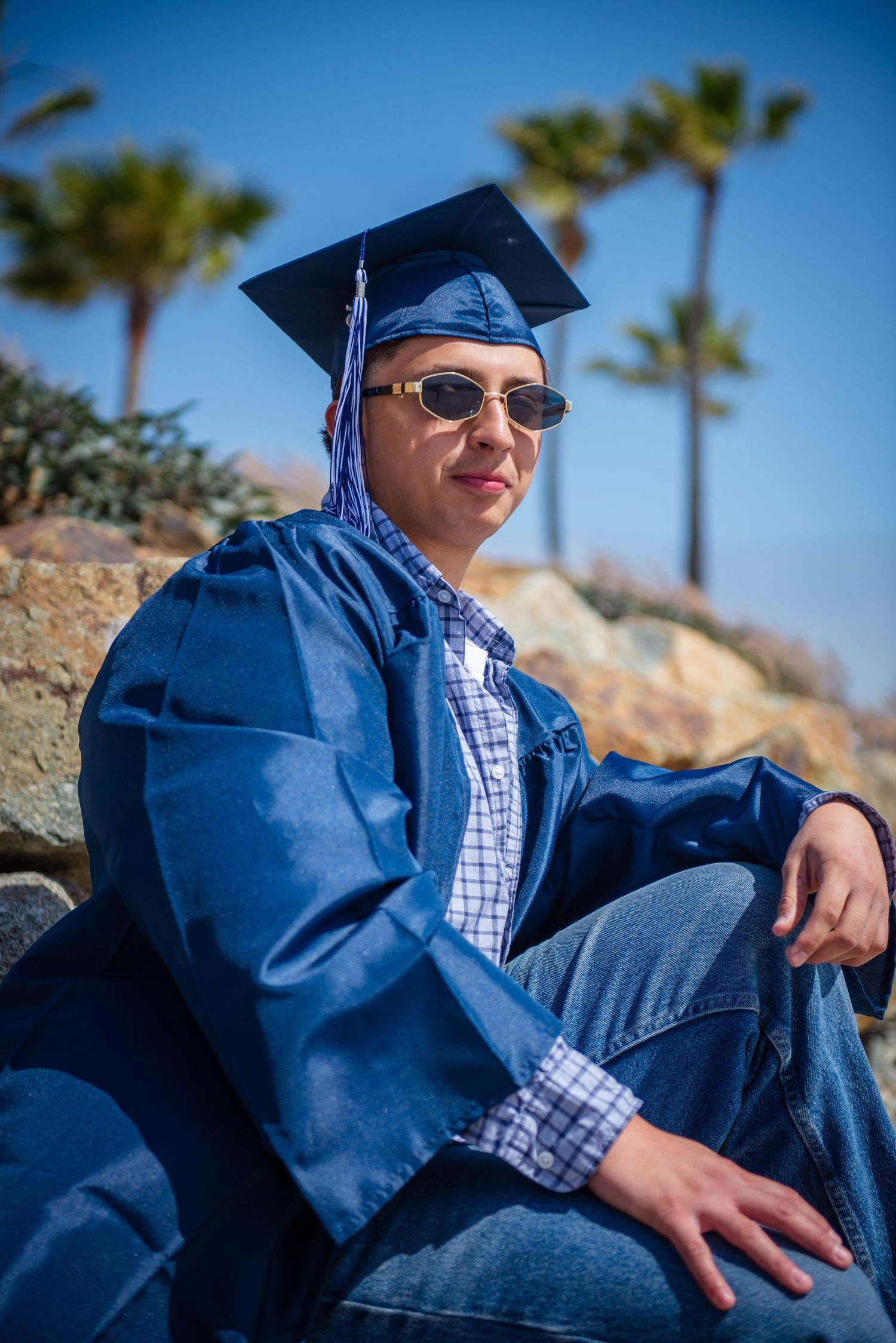 Graduate in blue cap and gown sits outdoors against palm tree backdrop wearing sunglasses on sunny day.